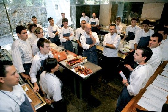 A large professional kitchen team listens to a briefing during a staff meeting.